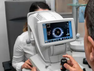 An eye doctor is using an ophthalmoscope to check a patient's eye while the patient sits and looks at the screen.