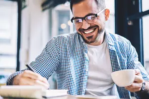 A smiling man wearing glasses is sitting in a restaurant and holding a cup of coffee while writing in a notebook