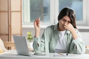 A woman sitting at a desk with a laptop and holding her glasses, looking frustrated.
