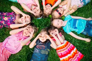 Group of young children lying on a lawn in a circle, smiling and making hand gestures