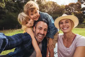 A family is smiling and taking a selfie in the park with a young girl on a man's shoulders and a boy on his back