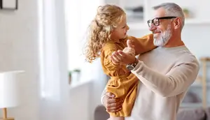 A smiling older man holds a young girl in his arms while dancing in a living room.