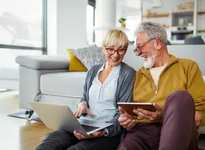 An elderly man and woman are sitting on the floor, looking at a laptop and tablet, and smiling.