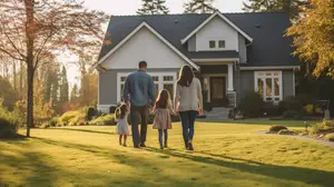 A family is walking in a grassy field in front of a white house with a gray roof, surrounded by trees and plants.