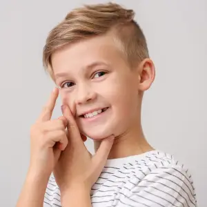 A smiling boy wearing a white striped shirt touching his cheek with his fingers