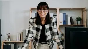 A smiling woman wearing glasses and a black and white checkered blazer standing in front of a bookshelf and a computer monitor.