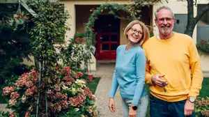 A smiling elderly couple stands outside their home with flowers and plants in the foreground.
