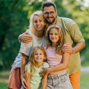 A family of four is smiling for a picture in a park with trees in the background