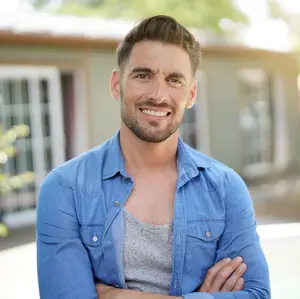 A smiling adult man with a beard and mustache wearing a blue shirt with his arms crossed in front of a house.