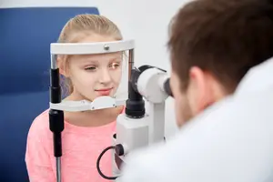 A girl with blonde hair is looking at the doctor with an ophthalmoscope on her head.