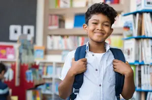 A smiling young boy with a backpack in a library with bookshelves and a blackboard in the background