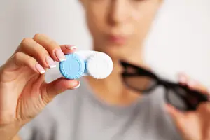 Woman holding a blue and white colored contact lens in one hand and a pair of sunglasses in the other.