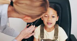A woman, likely a doctor, is examining a young girl's ear with a medical instrument, both are wearing casual clothing.