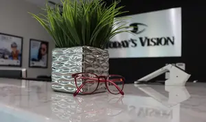 A pair of red-framed glasses rests on a counter, next to a silver vase with green plant inside. Behind the counter, a white sign reads 'Today's Vision'. On the wall, there are three framed images, and a device is placed on the right side of the counter.