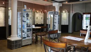 An interior view of an optical shop with chairs and tables arranged around display cabinets and frames, with lights hanging from the ceiling and a monitor on the wall.