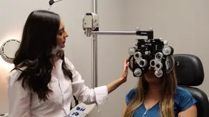 A woman wearing a white shirt is using an ophthalmoscope on a woman sitting on a chair in a clinic.