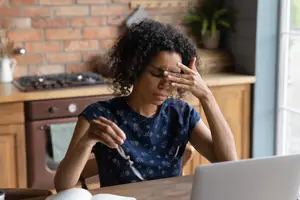 A woman is sitting at a desk in her kitchen, holding a pair of scissors and looking at a laptop with a serious expression, as if she is working on something.