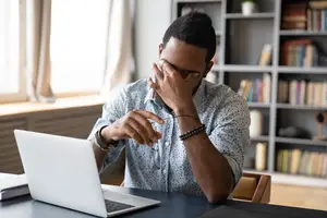A man in a room with a bookshelf is sitting at a desk and appears to be working on a laptop. He has a hand on his face and is holding a pair of glasses. There is a notebook and a cup on the desk. There is a window with curtains and a radiator behind him. 