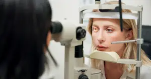 A woman in a white shirt having her eyes checked by a doctor with a phoropter.