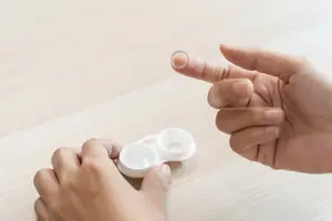 A person wearing a ring is holding a contact lens and a contact lens case on a wooden surface.