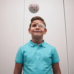 A boy wearing a turquoise shirt and eye patch smiles while looking at a ball with letters, against a plain background.