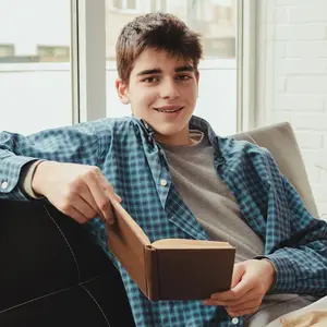 Smiling young person with braces reading a book on a sofa by a window.