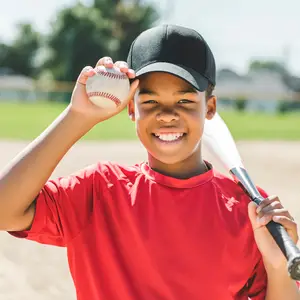 Smiling boy in a red shirt and black cap holding a baseball and bat on a sunny day.