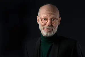 An older man with glasses, a beard, and a green turtleneck under a black coat, posing for a picture