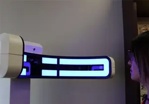 A woman wearing glasses is looking at a white and blue digital device with blue lights attached to a wall.