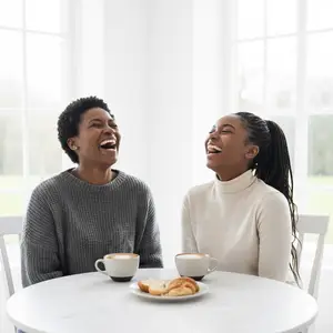 Two women sitting at a table and laughing with coffee and pastries in front of them.