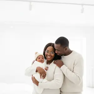A smiling couple and a baby are in a room, the baby is wrapped in a white blanket and is being held by the woman, while the man is standing behind them with his arm around the woman's shoulder. The room is white and clean, and there are lights on the ceiling.