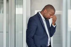 A man in a blue suit stands with his eyes closed and hand on his forehead in an office setting.
