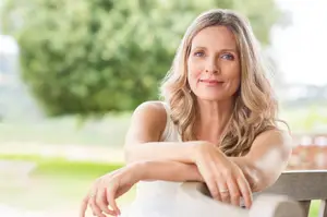 A smiling woman sitting on a bench in a park with her arms crossed.