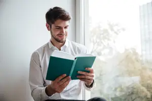 A man reading a book while sitting on a window sill