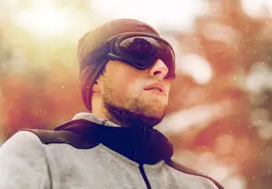 A man with a beard wearing a hat and goggles is looking up in the snow.