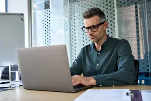 A man sitting on a chair while typing on a laptop on a table with papers, a glass of water, and a whiteboard on the side