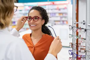 A woman is trying on glasses in a store while smiling and a woman adjusts them on her