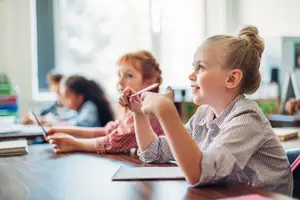 A girl holding a pencil points to her ear while sitting in a classroom with other students.