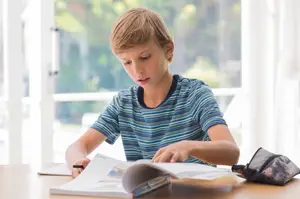 A boy is sitting on a chair in front of a desk with a book and a pouch on top, perhaps in a classroom, studying something from the book, and his head is slightly tilted to the side. Behind him is a glass window with a view of the outside, and the sunlight is shining through the window.