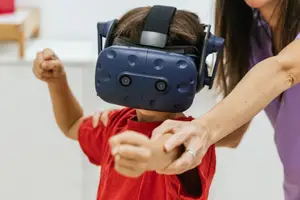 A young boy is wearing a blue virtual reality headset while a woman is holding his arm in a white room