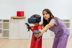 A woman in purple scrubs helps a child wearing a virtual reality headset and holding a controller in a room with white walls and wooden floors.