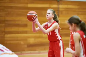 A girl holding a basketball and looking up, wearing a red jersey with white stripes and the number 11 on it, while another girl stands behind her, also wearing a red jersey, and both are playing basketball on a wooden court.