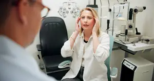 A woman sits in a chair with her hands on her head while looking at an eye doctor in an office.