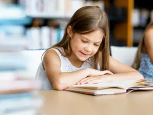 A girl sitting on a chair in a library reading a book and smiling
