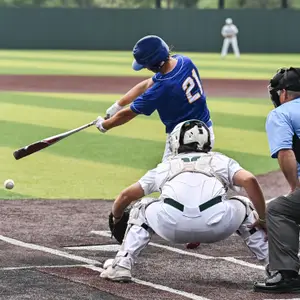 A baseball player is hitting the ball with a bat while another player is catching it in the foreground on a sunny day.