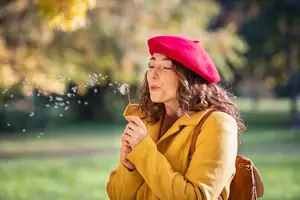 A woman wearing a red beret and yellow coat blowing on a dandelion
