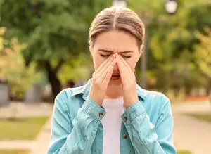A woman is standing on a pathway with her eyes closed and holding her face with both hands, perhaps sneezing