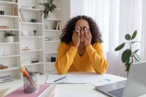 woman sitting at desk rubbing her eyes while working on a laptop