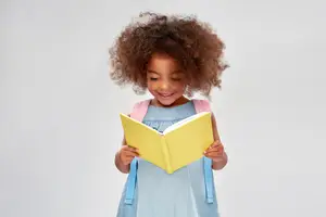 A young girl with curly hair is smiling while holding a yellow book and wearing a blue dress and a pink backpack.