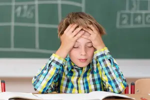 A young boy sitting in a classroom, holding his head with both hands and looking stressed.